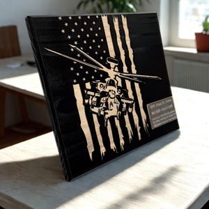May include: A wooden plaque featuring a laser-cut design of an American flag and a military helicopter. The flag's stripes are light-colored against a black background. A small metal plate with text is visible on the right side.