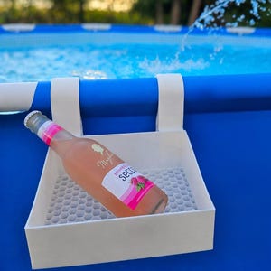 Könnte beinhalten: Ein weißer Kunststoff-Getränkehalter, der am Rand eines blauen Swimmingpools befestigt ist. Im Halter steht eine Glasflasche mit rosa Himbeer Secco, deren weißes Etikett ein Silhouetten-Profil und eine Abbildung von Himbeeren zeigt.