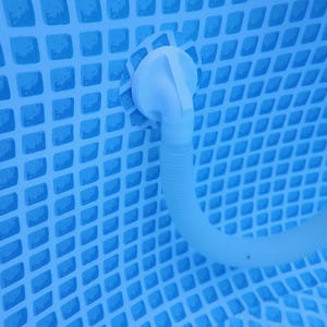 Könnte beinhalten: Eine weiße Pool-Einlassdüse aus Kunststoff und ein flexibler Wellschlauch, die an der Innenwand eines blauen Schwimmbeckens befestigt sind. Die Poolwand zeigt ein sich wiederholendes Gittermuster aus hellblauen Quadraten.