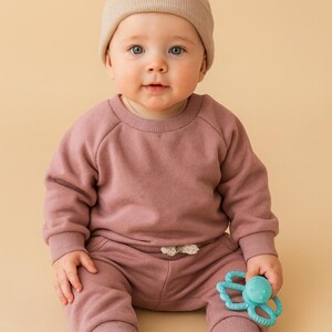 Może przedstawiać: Niemowlę w mauve'owej bluzie i spodniach dresowych, w czarno-białych trampkach i beżowej czapce. Dziecko trzyma niebieską zabawkę do gryzienia. Strój jest wygodny i swobodny.