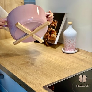 Könnte beinhalten: Ein hellrosa Keramiktopf wird über einem hölzernen Topfuntersetzer gehalten. Der Topf hat zwei Griffe und steht auf einer hölzernen Arbeitsplatte. Eine weiß-rosa gesprenkelte Flasche steht auf einem weißen Untersetzer. Ein Kochbuch ist im Hintergrund.