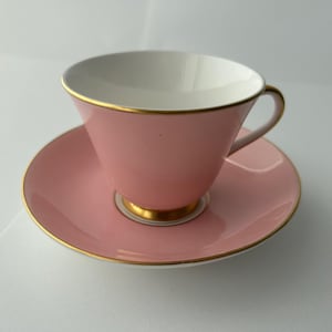 May include: A pink teacup and saucer set. The teacup and saucer are a soft pink color with gold trim. The inside of the teacup is white. The set is likely made of porcelain or a similar material.