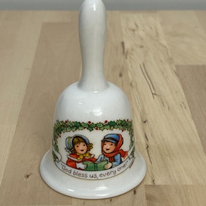 Puede incluir: Campana de cerámica blanca con un mango largo y una base acampanada. La campana presenta una ilustración colorida de dos niños y el texto "God bless us, every one!" rodeado por una guirnalda verde de acebo.