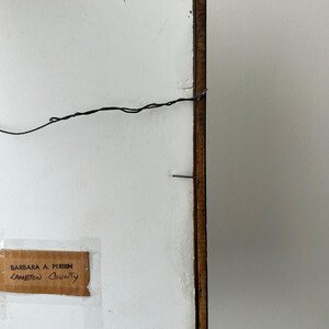 May include: Back of a framed artwork with a brown wooden frame. The artwork's backing is white with a black wire for hanging. A label with the text "BARBARA A. PERRIN" and "LAMBTON County" is attached.