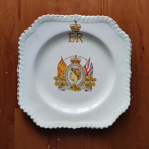 Op de afbeelding: Een vierkant wit keramieken bord met een geschulpte zilveren rand. Het midden toont een portret van Koningin Elizabeth II, de tekst Coronation June 2nd 1953, twee vlaggen en bloemmotieven. Bovenaan staat de gouden tekst E II R onder een koninklijke kroon.