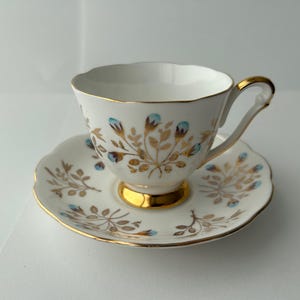 May include: White teacup and saucer set with gold trim and floral design. The teacup has a gold handle and base. The saucer has a scalloped edge. The floral design includes gold and blue accents.