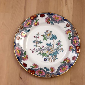 May include: A round, white ceramic plate with a gold rim. The plate features a floral design with pink, blue, and yellow flowers. The border has a decorative pattern in blue, gray, and gold. The plate is set on a light brown wooden surface.