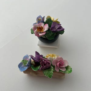 Puede incluir: Dos arreglos florales de cerámica. Uno es un pequeño cuenco negro lleno de flores rosas, moradas, amarillas y azules. El otro es una jardinera rectangular con flores similares. Hojas verdes acentúan las coloridas flores.