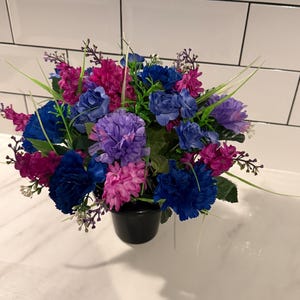 May include: A vibrant floral arrangement in a black pot. The bouquet features a mix of blue, purple, and pink artificial flowers with green foliage. The arrangement is set against a white tiled background.