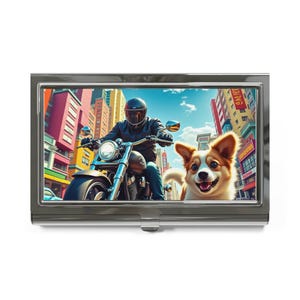 May include: A silver business card holder featuring a vibrant illustration of a person riding a motorcycle through a city, with a smiling corgi dog in the foreground. The background includes colorful buildings and a blue sky.