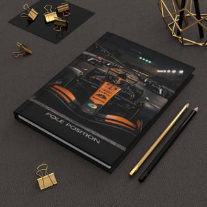 Puede incluir: Diario negro con una foto de un coche de carreras de Fórmula 1 en naranja y negro. La portada tiene las palabras "POLE POSITION" impresas. También se ven lápices dorados y negros y clips dorados.