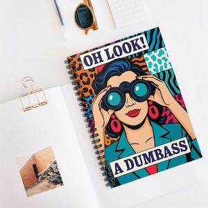 May include: Spiral-bound notebook with a pop art illustration of a woman looking through binoculars. The cover features the text "OH LOOK! A DUMBASS" in a bold font, set against a colorful background with leopard print and geometric patterns.