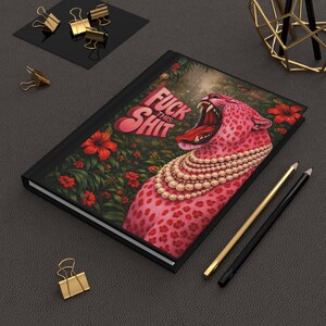 May include: Black hardcover journal featuring a pink leopard with a pearl necklace and the text "FUCK THIS SHIT." The leopard is set against a floral background with red flowers. Gold and black pencils are on the side.