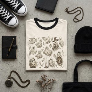 May include: A cream-colored ringer t-shirt with a black collar and sleeve trim. The center graphic features a detailed, vintage-style illustration of multiple ghosts, small pumpkins, a lit candle, and botanical sprigs in shades of brown and gray.