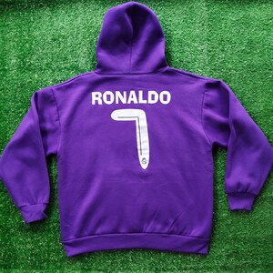 May include: A purple hooded sweatshirt with the name "RONALDO" printed in white across the upper back. Below the name is a large white number "7" with a silver stripe and a small emblem at the bottom. The sweatshirt has a hood and long sleeves.