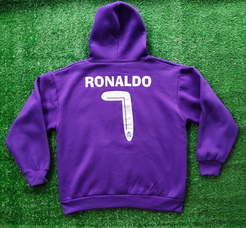 May include: A purple hooded sweatshirt with the name "RONALDO" printed in white across the upper back. Below the name is a large white number "7" with a silver stripe and a small emblem at the bottom. The sweatshirt has a hood and long sleeves.