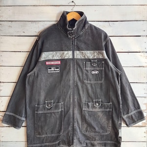 May include: A charcoal gray denim jacket with a high collar and a full-length zipper. The jacket features two chest pockets, two lower pockets, and a silver stripe across the chest. Patches on the chest read "WORLD CHAMPIONSHIP" and "BFT RACING TEAM".