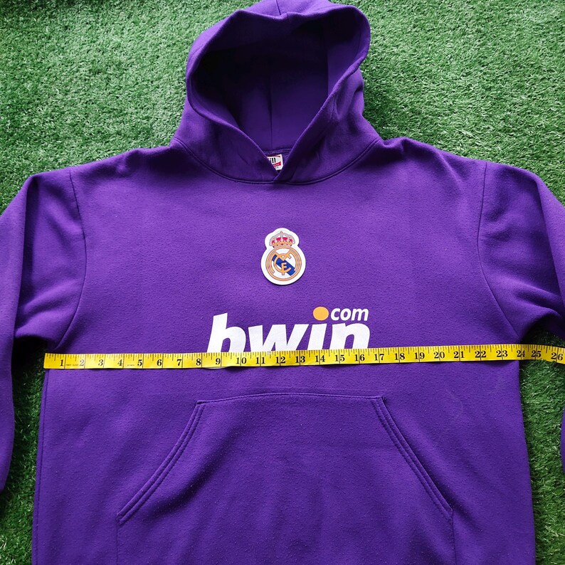May include: A purple hoodie with a front pocket and a hood. The hoodie features a team logo and the text "hwin .com". The hoodie is measured with a tape measure, showing a width of approximately 26 inches.