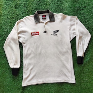 May include: Cream-colored long-sleeve rugby shirt with a gray collar and black cuffs. Features the "Steinlager" logo, a Canterbury logo, and the "All Blacks" logo with a silver fern.