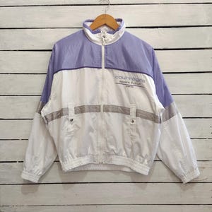 May include: A white and light purple zip-up windbreaker jacket featuring a high collar, elastic cuffs, and two front pockets. The chest displays the text courr&egrave;ges sport futur paris in dark lettering, with a horizontal grey stripe across the midsection.