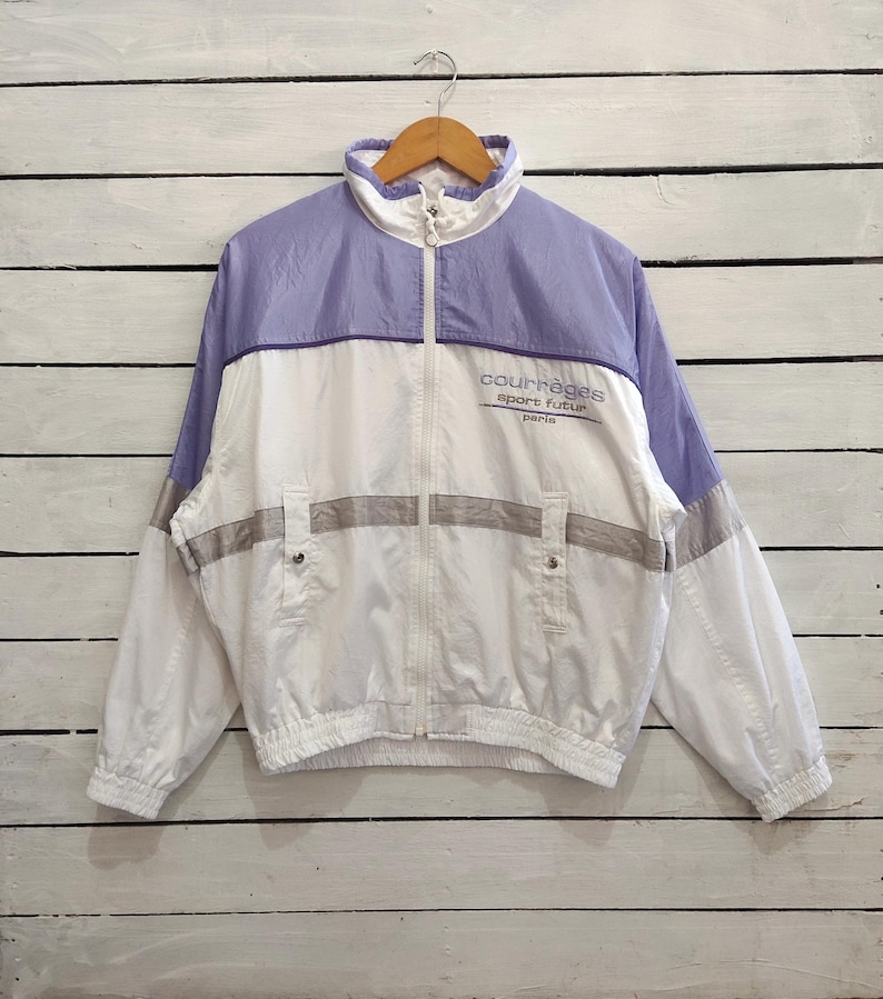 May include: A white and light purple zip-up windbreaker jacket featuring a high collar, elastic cuffs, and two front pockets. The chest displays the text courr&egrave;ges sport futur paris in dark lettering, with a horizontal grey stripe across the midsection.