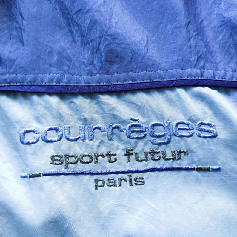 May include: A light blue nylon garment featuring a dark blue horizontal seam at the top. The fabric displays embroidered text in dark blue and silver thread that reads courr&egrave;ges sport futur paris with a horizontal line separating the words.
