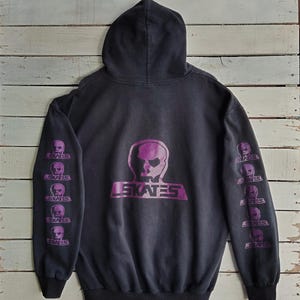 Puede incluir: Sudadera con capucha negra con un diseño de calavera morada y el texto "LE SKATES". Las mangas muestran gráficos de calaveras moradas repetidos. La sudadera con capucha está hecha de un material suave y cómodo.