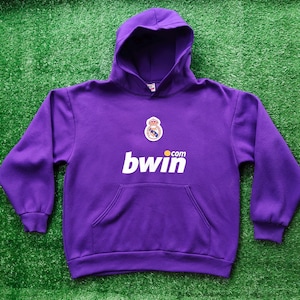 May include: A purple hooded sweatshirt with a front pocket and a white "bwin.com" logo. The sweatshirt features a team crest and is set against a green background.