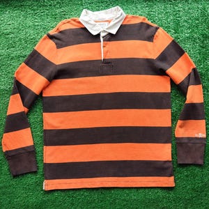 Op de afbeelding: Een rugby shirt met lange mouwen en horizontale strepen in donkerbruin en feloranje. Het shirt heeft een witte kraag, een knoopsluiting met witte bies en een klein wit geborduurd logo van een schedel met gekruiste botten op de linkermanchet.