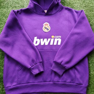 May include: A purple hooded sweatshirt with a front pocket and ribbed cuffs. The sweatshirt features a team crest and the word "bwin" in white block letters. The garment is made of a soft, textured fabric.