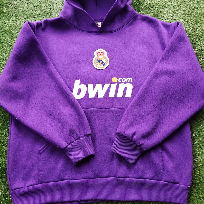 May include: A purple hooded sweatshirt with a front pocket and ribbed cuffs. The sweatshirt features a team crest and the word "bwin" in white block letters. The garment is made of a soft, textured fabric.