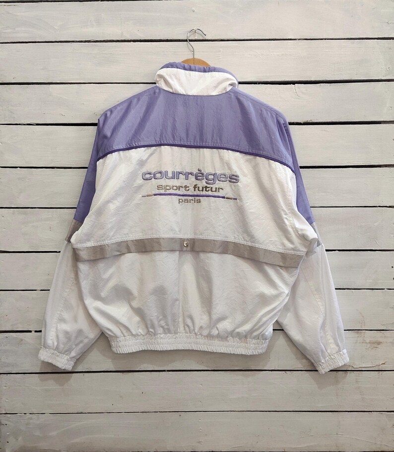 May include: A white and light purple windbreaker jacket featuring a high collar, long sleeves with elastic cuffs, and a cinched elastic hem. The back displays embroidered text reading courr&egrave;ges sport futur paris above a horizontal grey stripe.