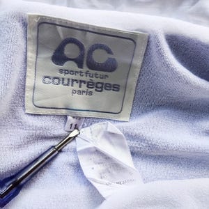 May include: A light purple garment featuring a white rectangular label with the text AC sport futur courr&egrave;ges paris. A smaller white tag below displays the number 11. A blue and silver tool points toward the tags on the textured fabric.