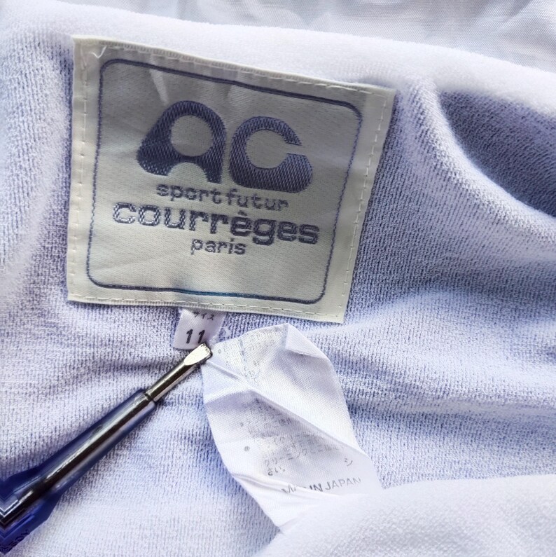 May include: A light purple garment featuring a white rectangular label with the text AC sport futur courr&egrave;ges paris. A smaller white tag below displays the number 11. A blue and silver tool points toward the tags on the textured fabric.