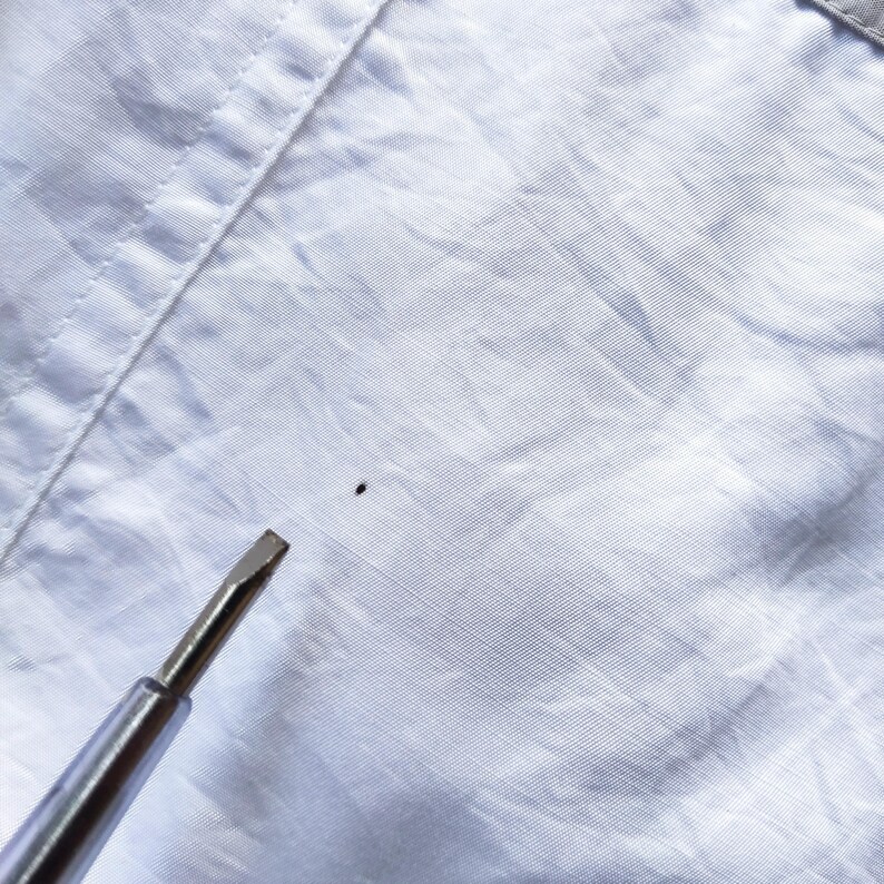 May include: A light blue button-down shirt featuring a visible small dark spot on the fabric surface. A metal screwdriver tip is positioned near the spot to indicate its location on the textured, woven material of the garment.