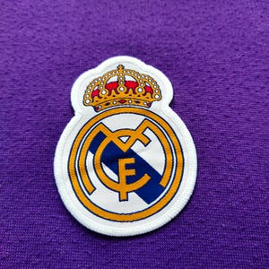 May include: A white patch featuring the Real Madrid crest, with a gold crown, a gold and blue circular emblem, and the letters "MCF". The patch is on a purple fabric background.