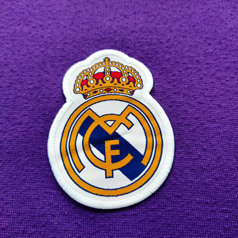 May include: A white patch featuring the Real Madrid crest, with a gold crown, a gold and blue circular emblem, and the letters "MCF". The patch is on a purple fabric background.