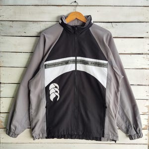 May include: A gray, black, and white zip-up jacket with a high collar. The jacket features a white stripe across the chest with text and a white logo on the front. The sleeves and upper back are gray.