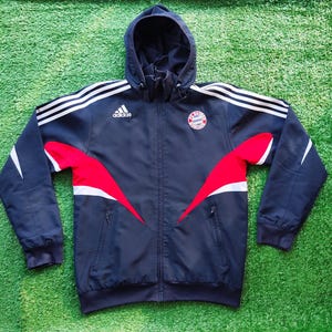 Può includere: Una giacca con cappuccio e zip blu navy scuro con il logo Adidas bianco sul petto destro e lo stemma del FC Bayern Monaco sul petto sinistro. La giacca presenta strisce bianche sulle spalle e dettagli curvi di colore rosso e bianco.