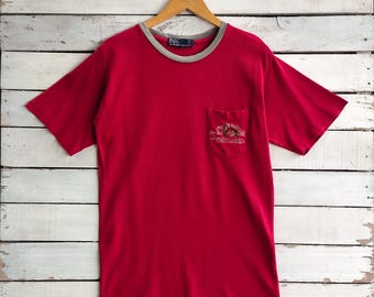 May include: A red t-shirt with a gray trim around the collar and sleeves, and a small pocket on the chest. The pocket features an embroidered design. The shirt is hanging on a wooden hanger.