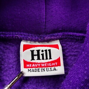 May include: Close-up of a purple hoodie with a tag that reads "Hill." The tag has a red and white design with the words "Heavy Weight" and "Made in U.S.A." printed on it. The hoodie is made of a thick, textured material.