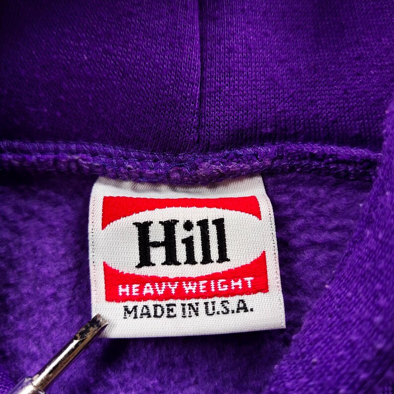 May include: Close-up of a purple hoodie with a tag that reads "Hill." The tag has a red and white design with the words "Heavy Weight" and "Made in U.S.A." printed on it. The hoodie is made of a thick, textured material.