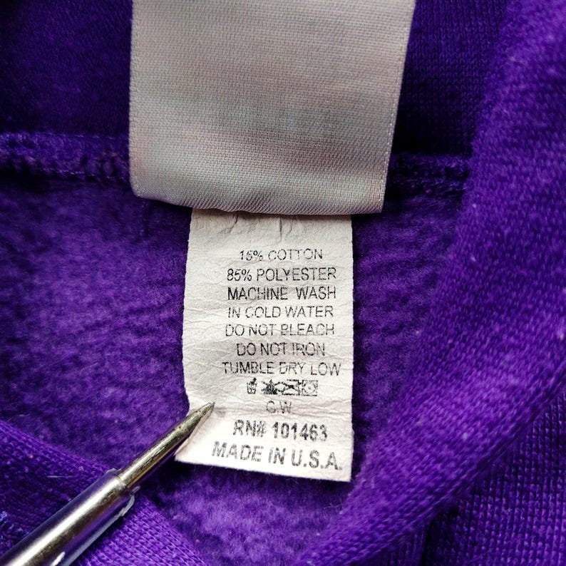 May include: Close-up of a purple fabric with a white tag attached. The tag includes washing instructions: 15% cotton, 85% polyester, machine wash in cold water, do not bleach, do not iron, tumble dry low. The tag also reads "MADE IN U.S.A."