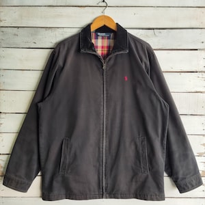 以下が含まれることがあります： フルジップとコーデュロイの襟が付いた濃いグレーのジャケット。襟の内側は赤、黄、青のチェック柄。胸には小さな赤いロゴがあります。