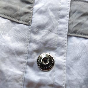 May include: A white garment featuring a central vertical placket with visible stitching and grey fabric panels on the sides. A circular silver-toned metal snap button is centered on the placket, embossed with the text Courr&egrave;ges Paris.