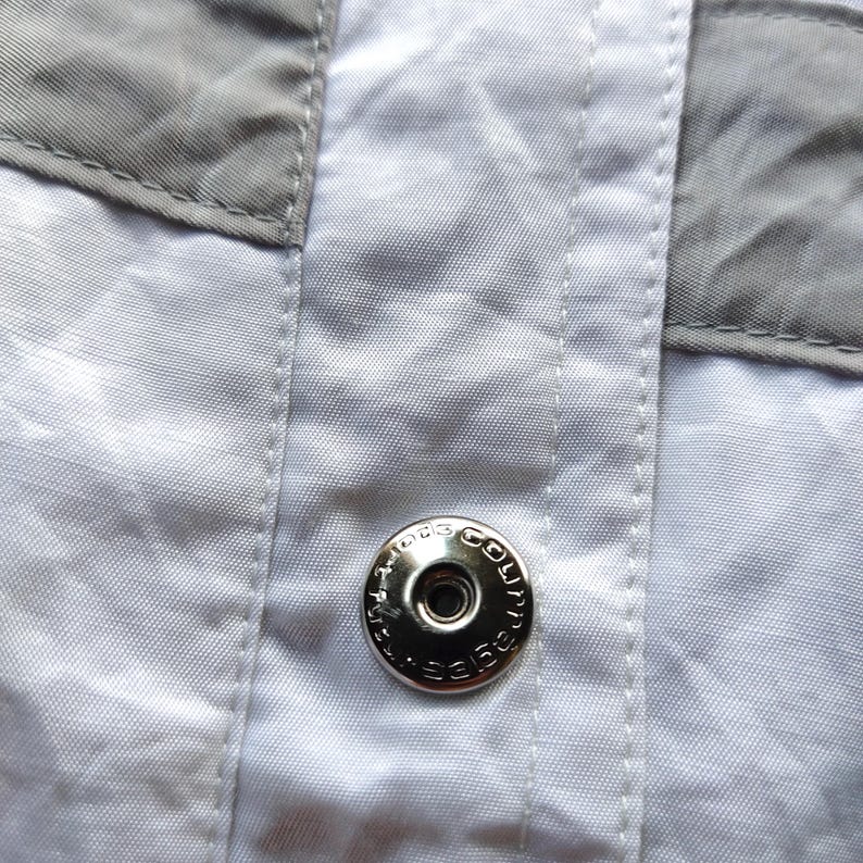 May include: A white garment featuring a central vertical placket with visible stitching and grey fabric panels on the sides. A circular silver-toned metal snap button is centered on the placket, embossed with the text Courr&egrave;ges Paris.