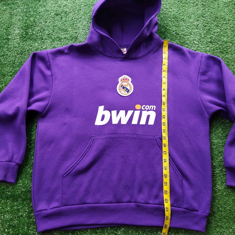 May include: A purple hooded sweatshirt with a front pocket and a drawstring hood. The sweatshirt features a crest and the word "bwin" in white. A yellow measuring tape is placed on the right side of the sweatshirt.