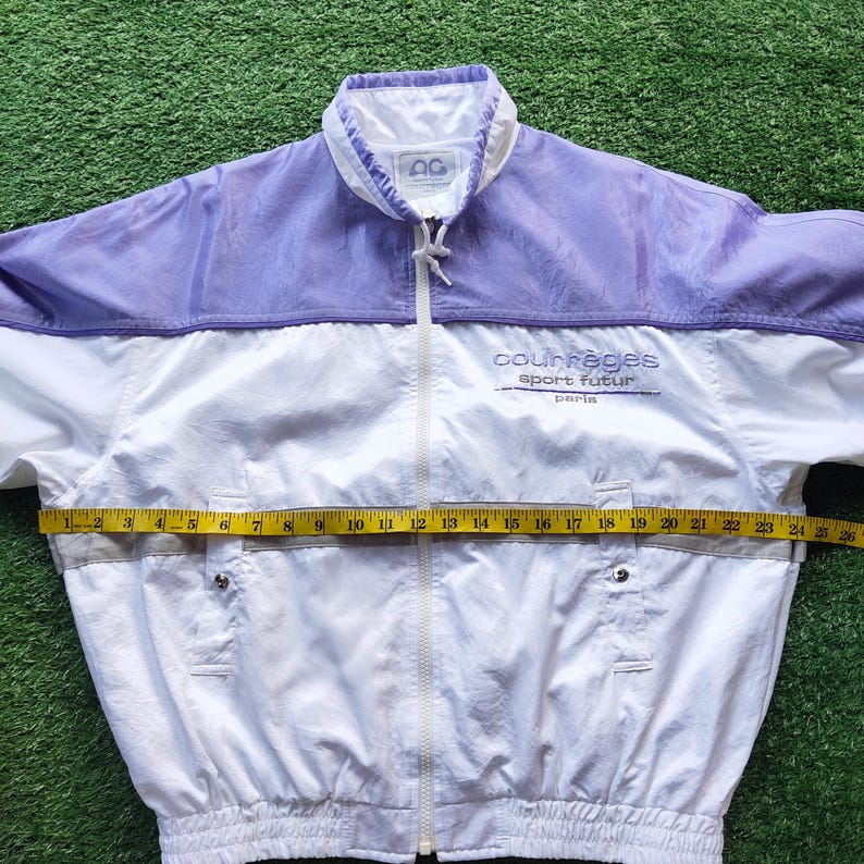 May include: A white and light purple windbreaker jacket with a full-length front zipper, a high collar, and two front pockets. The chest features the text 'courr&egrave;ges sport futur paris'. A yellow measuring tape shows a width of 26 inches.