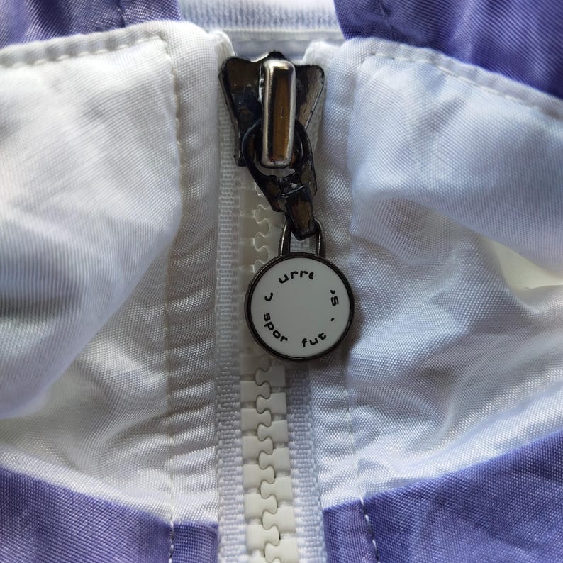 May include: A white plastic zipper closure on a light purple and white garment. The metal zipper pull features a circular white charm with the black text "curre s spor fut" printed around the edge, attached to a silver-toned metal slider.