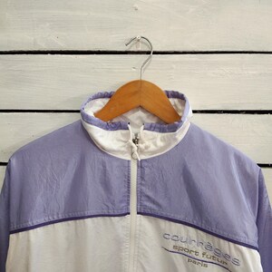 May include: A light purple and white windbreaker jacket featuring a high collar with a front zipper closure. The chest area displays the embroidered text "courr&egrave;ges sport futur paris" in a dark purple thread on the white lower section.